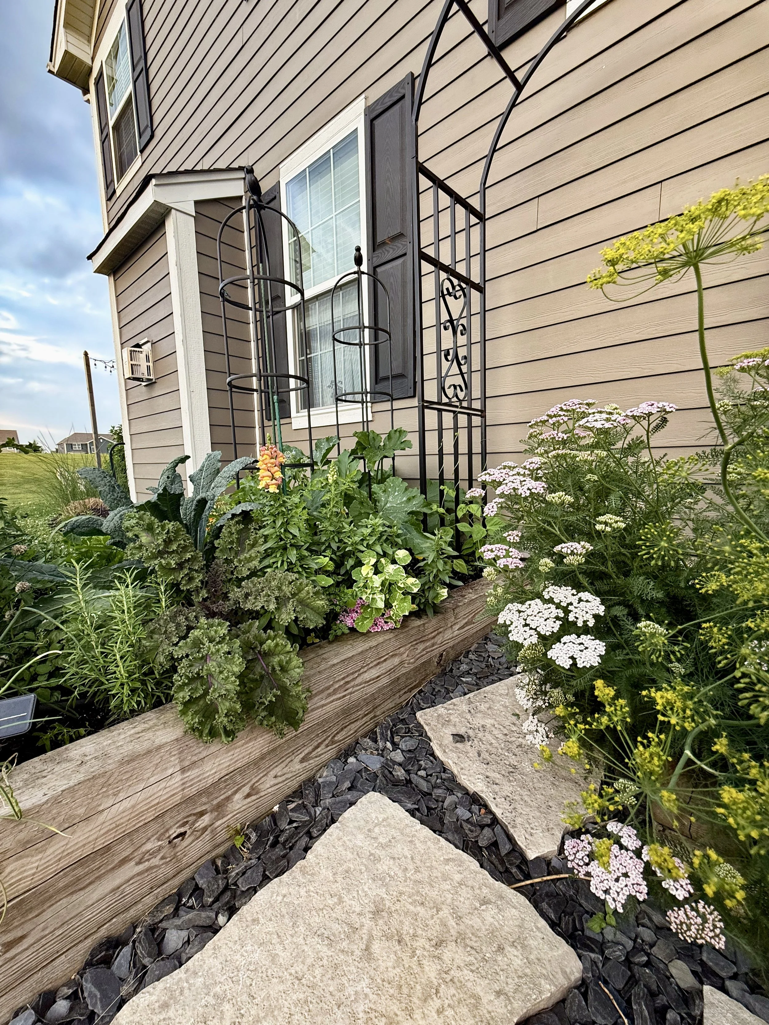 A close-up of a garden bed with various plants and flowers next to a stone and wood pathway in front of a house with beige siding, a window with black shutters, and a small metal trellis.