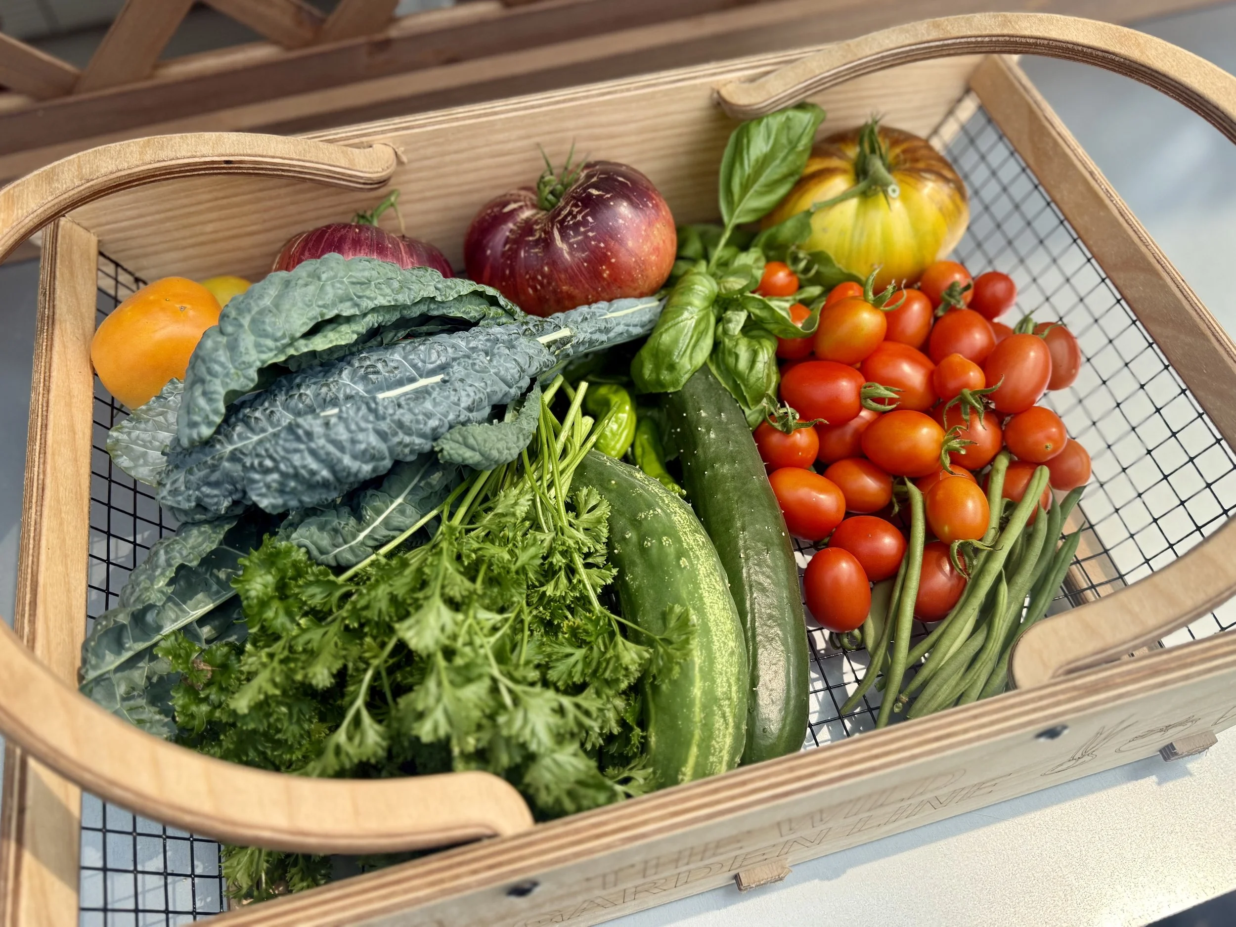 A wooden basket contains fresh vegetables including cherry tomatoes, heirloom tomatoes, cucumber, kale, basil, and other greens.