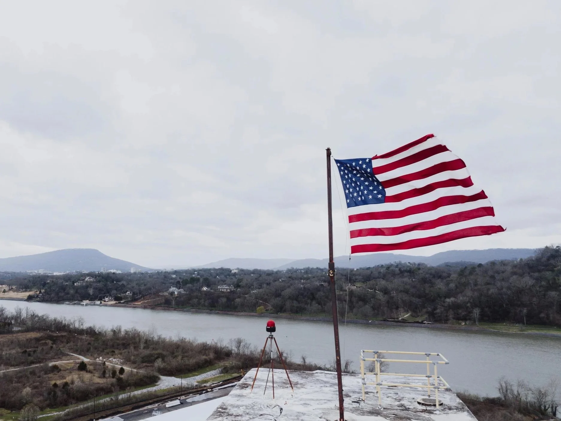 American flag flying on a pole overlooking a river, with mountains and a cloudy sky in the background.