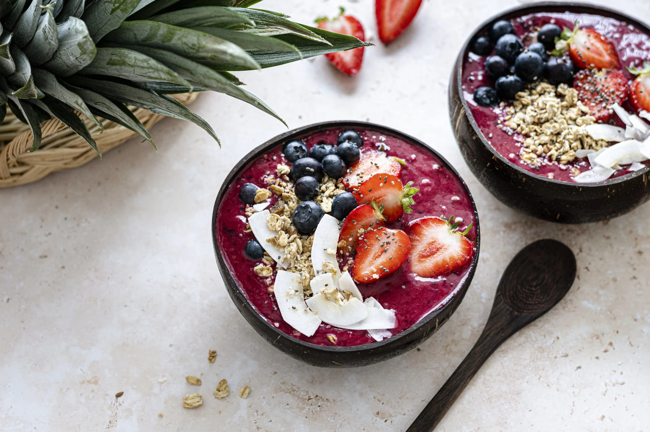 Two bowls of smoothie topped with strawberries, blueberries, oats, and coconut flakes, placed on a white surface with a pineapple and a wooden spoon nearby.