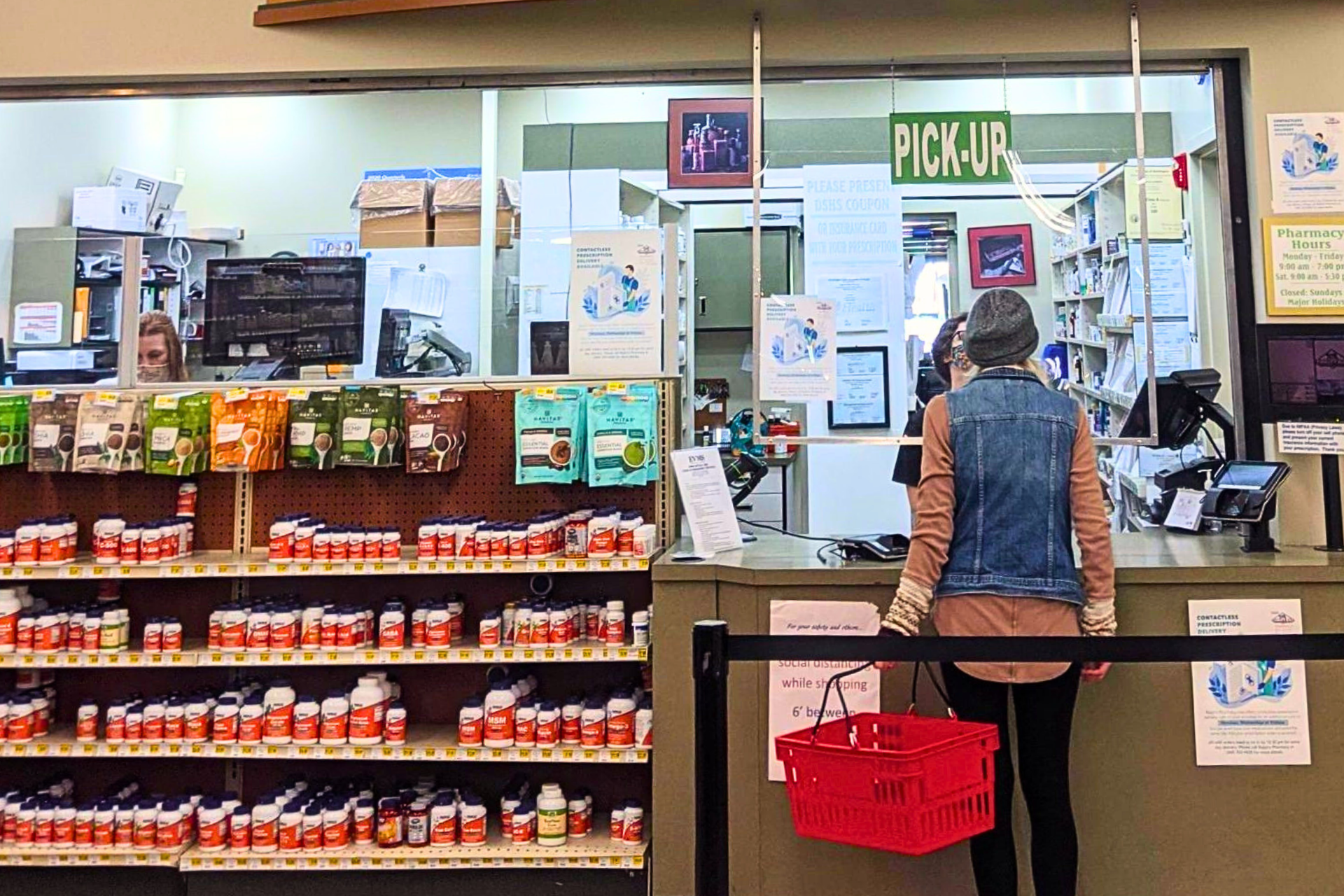 Customer at pharmacy pickup counter with shelves of medicine bottles and supplements in the foreground, and a sign indicating 'PICK-UP' at the counter.