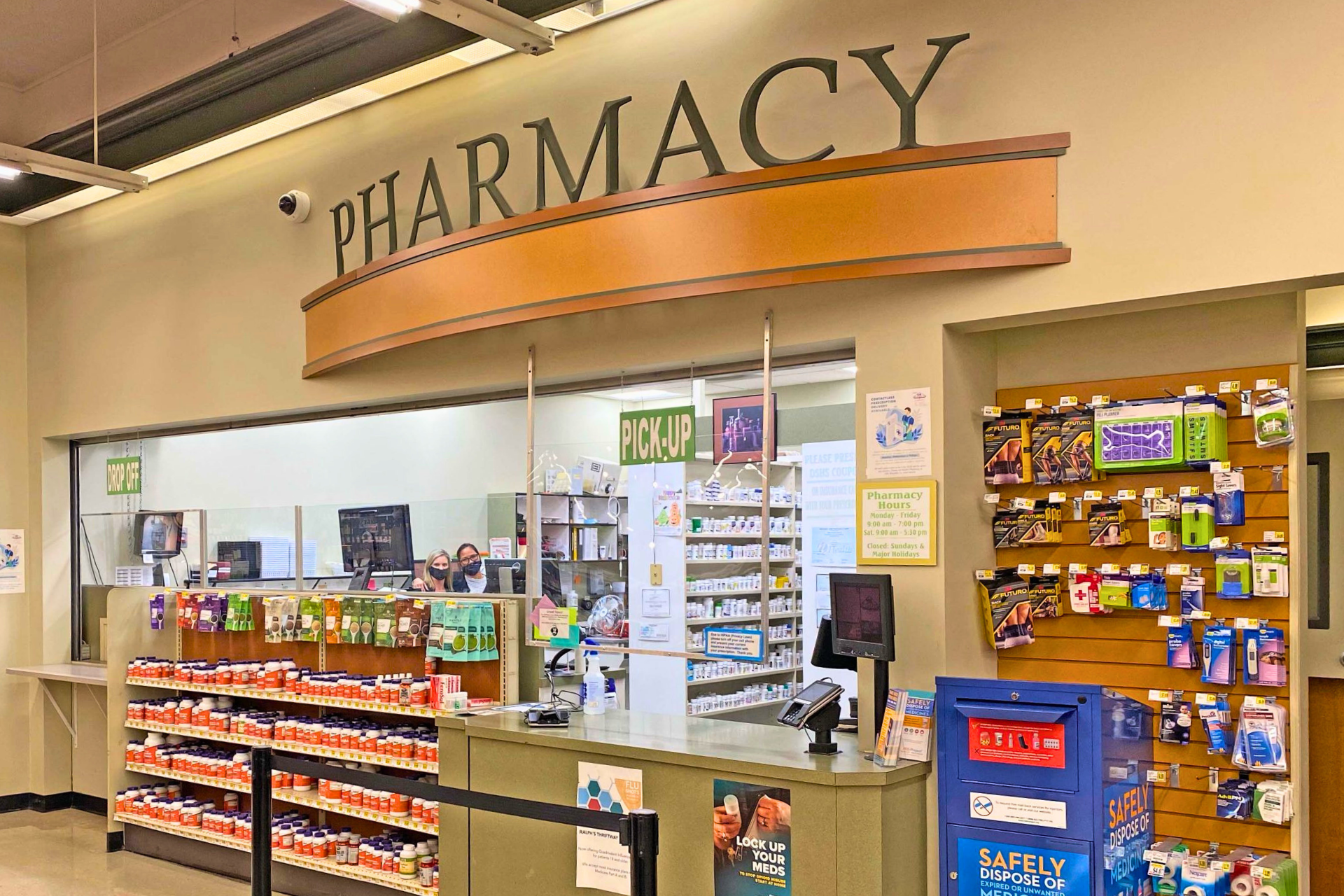 Inside a pharmacy with a counter, shelves stocked with medication or health products, and two staff members wearing masks behind the counter. There's signage indicating 'Drop Off', 'Pick Up', and pharmacy hours displayed.