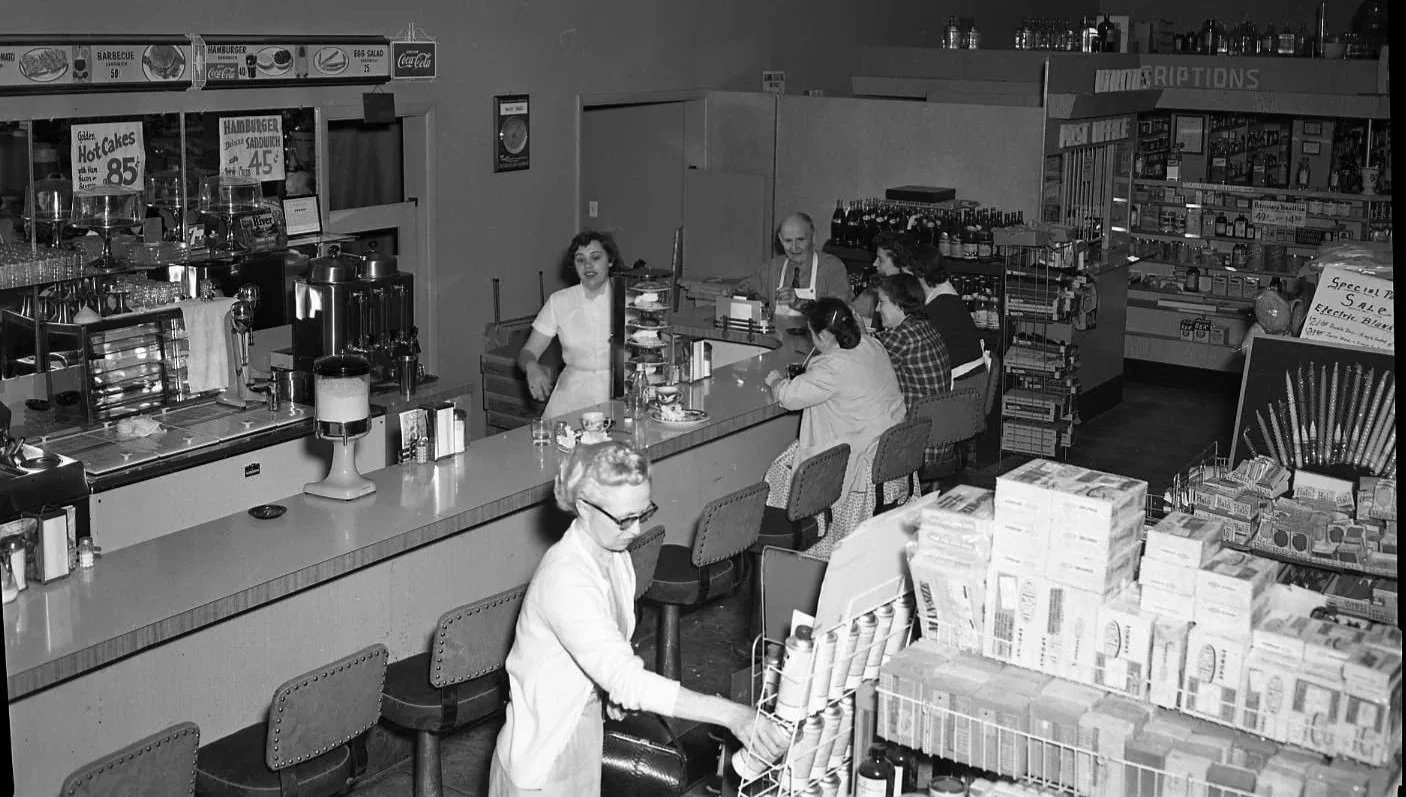 Black and white photo of a vintage soda fountain and grocery store with a counter, several women and a man sitting and working, and a waitress behind the counter serving customers.