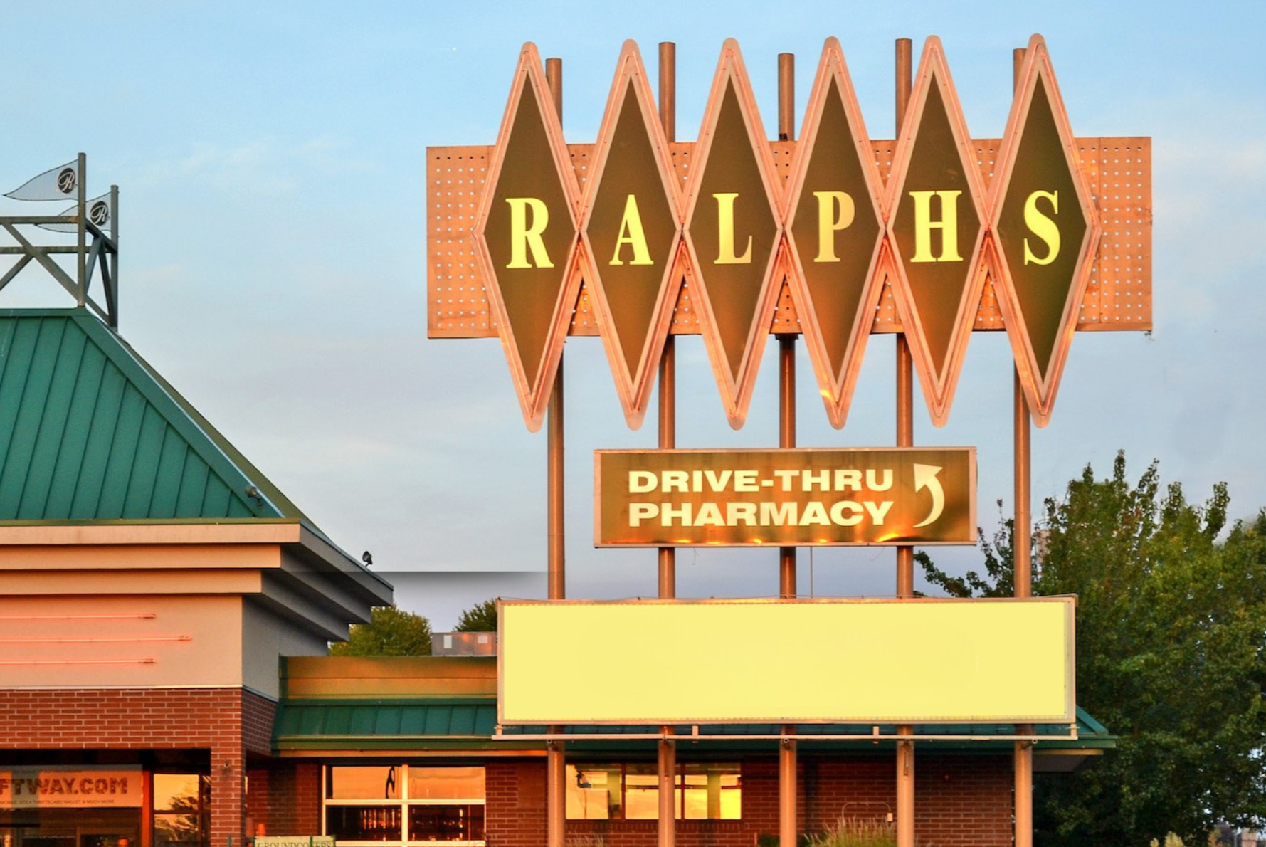 Ralph's store sign with a large diamond-shaped logo overhead and a smaller sign below advertising drive-thru pharmacy, with green roofing and brick facade, trees in the background, and a clear sky.