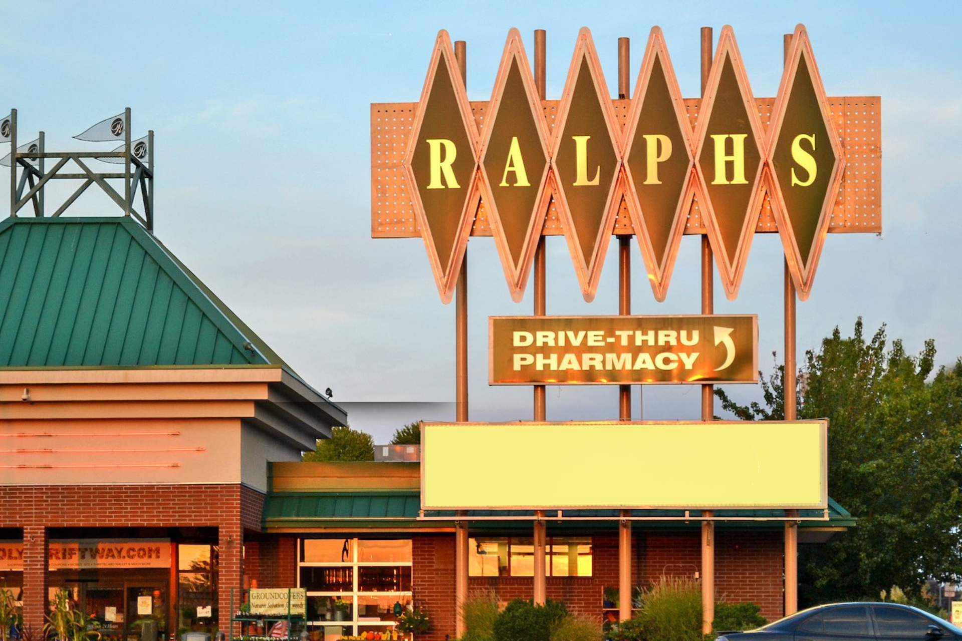 Sign for Ralphs grocery store with drive-thru pharmacy underneath, billboards, and store entrance with plants and displays.