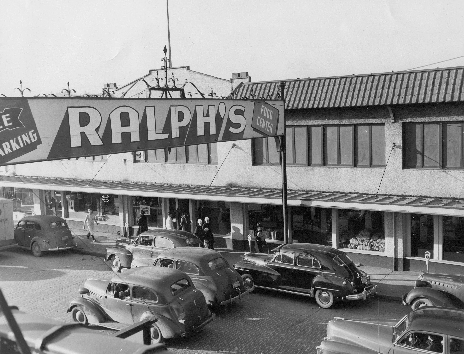 Black and white photograph of an outdoor shopping area with cars parked in front and people walking on the sidewalk. A large sign reads 'RALPH'S FOOD CENTER', and there are storefronts with large windows on the building behind the parked cars.