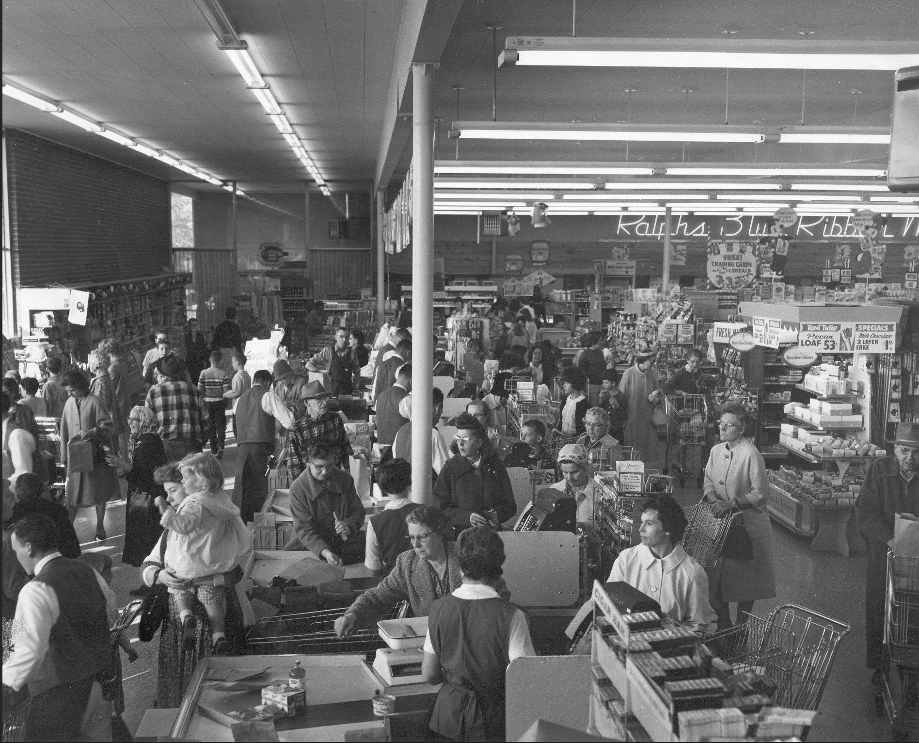 A busy scene inside a grocery store or supermarket with people shopping, checking out, and browsing shelves. The scene is in black and white.