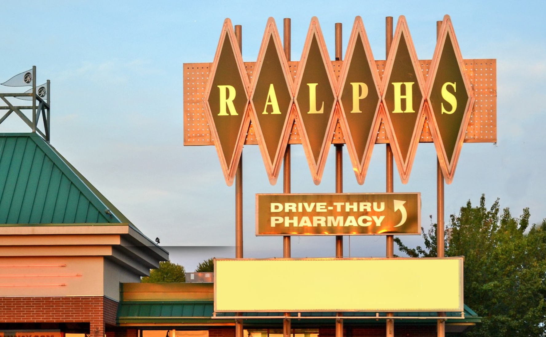 A large vintage-style sign for Ralphs grocery store with a bright, illuminated design. The sign reads "RALPHS" and includes a smaller sign below that says "DRIVE-THRU PHARMACY" with an arrow indicating the direction. The background sky is partly cloudy, and there are trees and part of the store building visible.