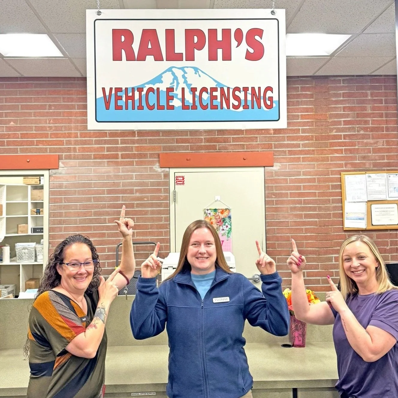 Three women standing inside a building beneath a sign that reads 'Ralph's Vehicle Licensing' with a mountain graphic. The women are smiling and pointing upwards at the sign.
