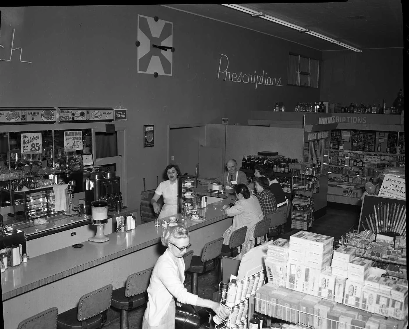 A vintage grocery store or supermarket scene with a counter serving food, a woman behind the counter, and several customers sitting or shopping, with shelves and products in the background.
