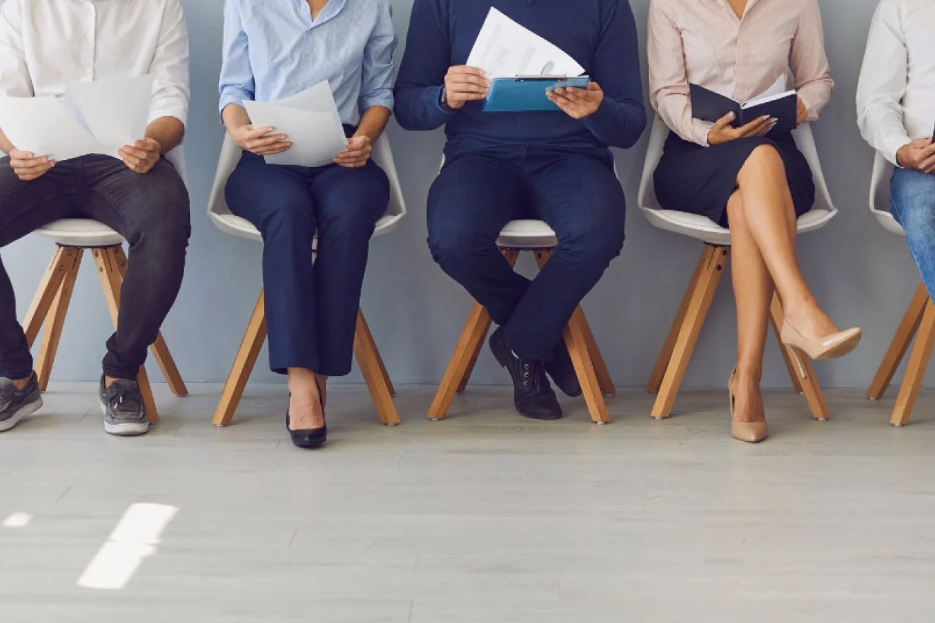People sitting in a row on chairs, holding documents or tablets, waiting.