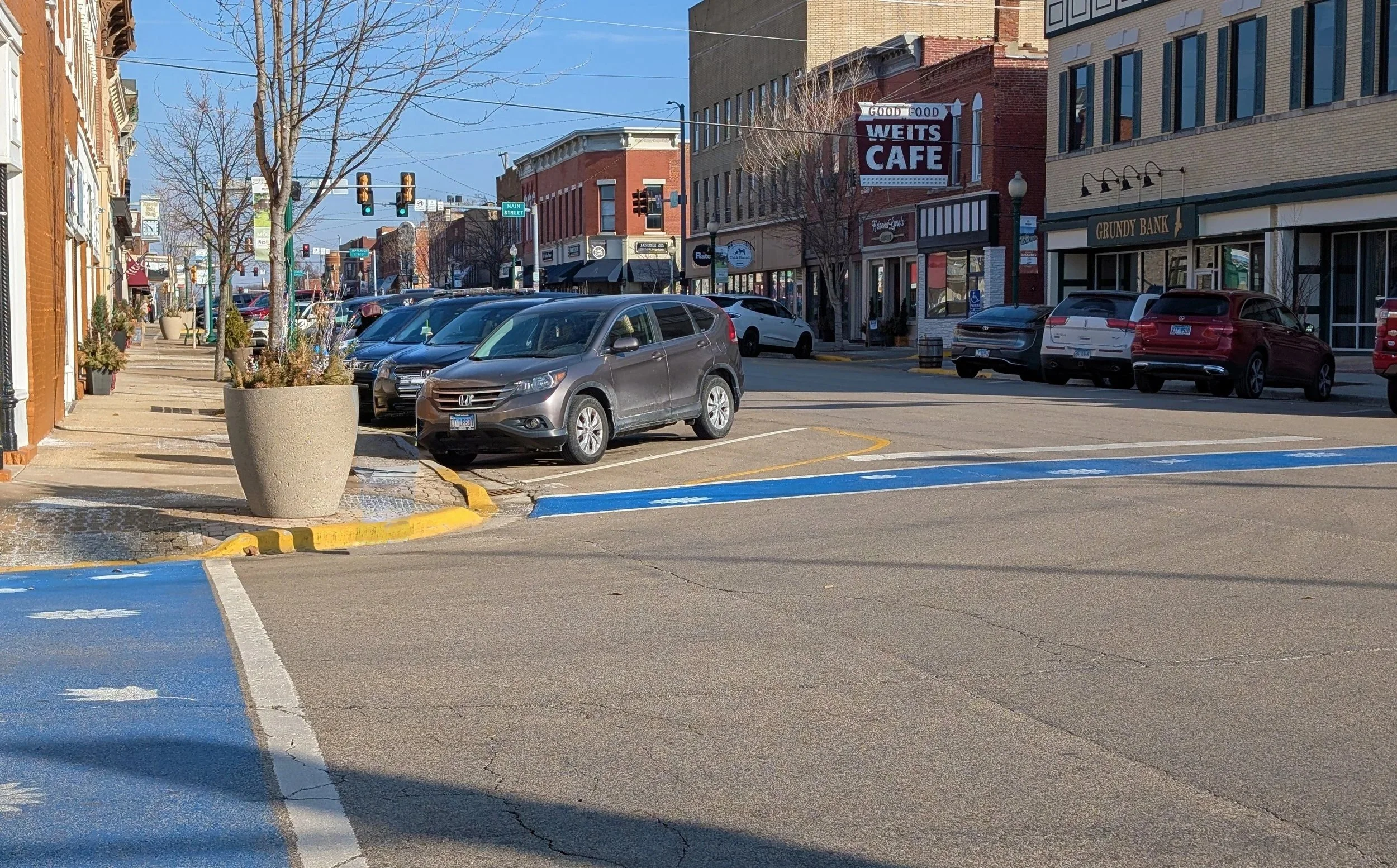 A downtown street scene with parked cars along the curb, storefronts, leafless trees, and a traffic light at an intersection on a clear day.