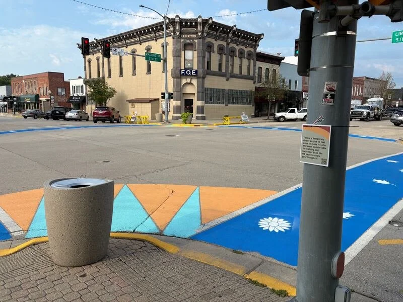 Street corner with a colorful bike lane, a sidewalk, a trash can, a pole with a sign, and a historic brick building with a sign 'F.O.E.' in a small town.