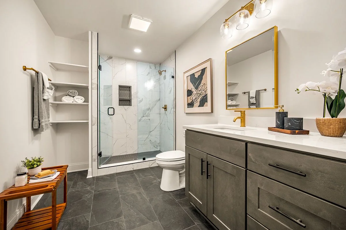 Modern bathroom with shower enclosure, gray cabinetry, white countertop, gold fixtures, and minimal decor.