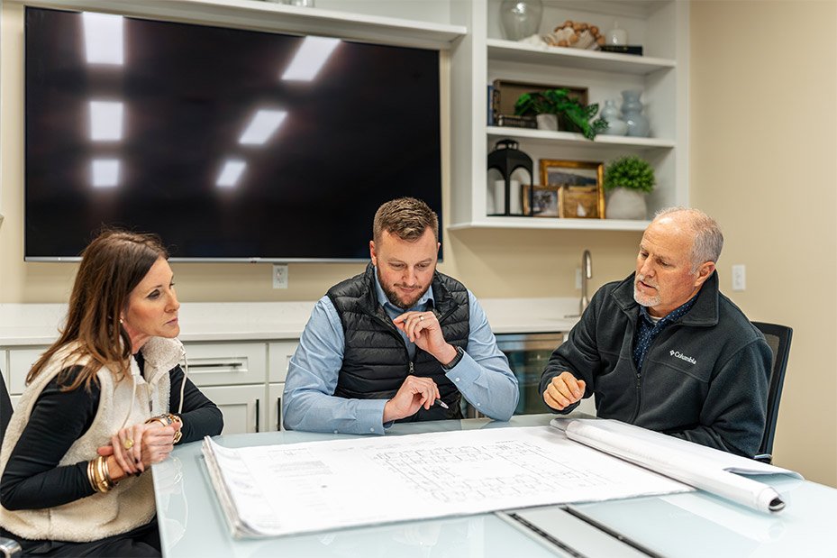 Three people seated at a table reviewing large blueprints in an office conference room.