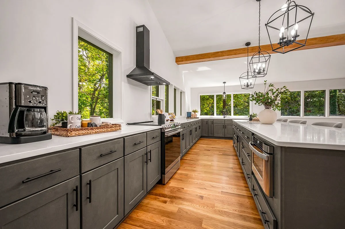 Modern kitchen with gray cabinets, white countertops, and large windows showing green trees outside. There is a coffee maker, a basket with cups, and a potted plant on the counter. Three decorative hanging light fixtures are above the island, which has a flower vase.