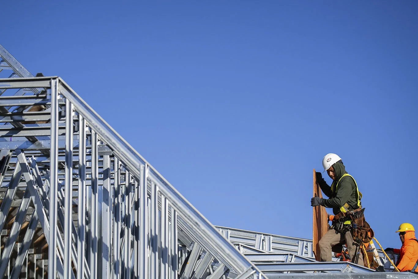 Construction workers installing metal panels on a roof under a clear blue sky.