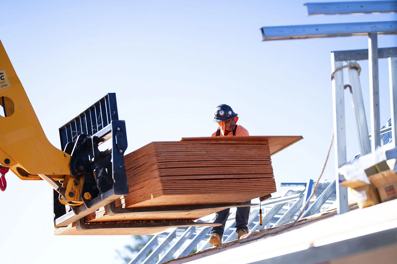 Construction worker operating a crane to lift a stack of plywood sheets on a construction site.