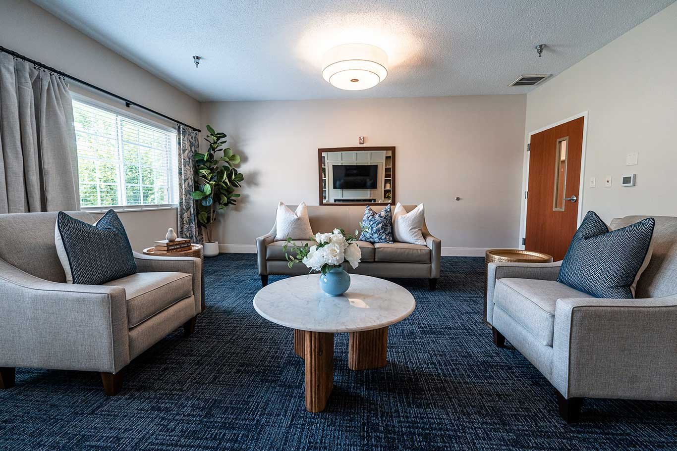 Living room with three beige sofas, dark carpet, round marble coffee table with a vase of white flowers, large window with curtains, potted plant, wall-mounted TV, wooden door.