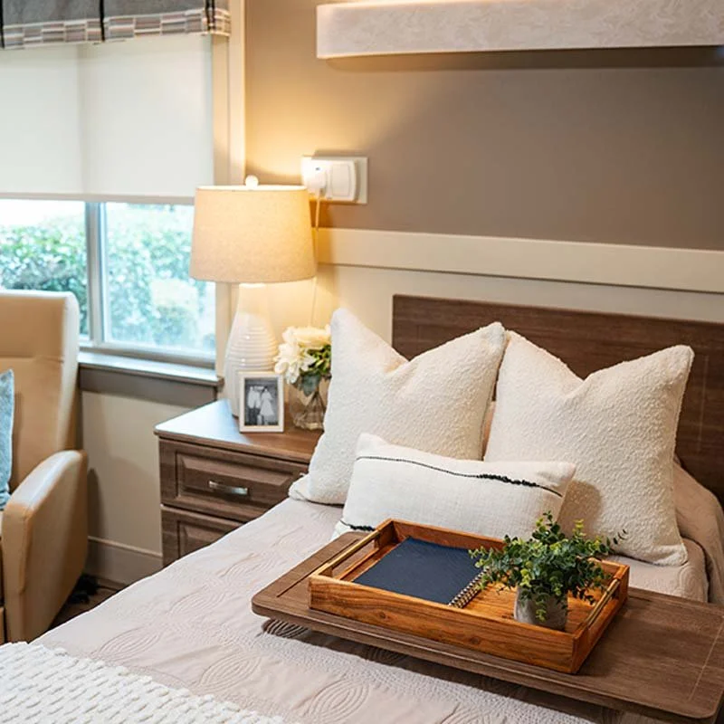 A cozy bedroom corner with a neatly made bed featuring multiple pillows, a wooden tray holding a notebook and a potted plant, a bedside table with a lamp, framed photo, and flowers, and a window with blinds allowing natural light.