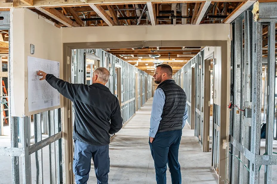 Two men inside a building under construction, discussing a blueprint on the wall, with exposed metal wall studs and unfinished ceiling.
