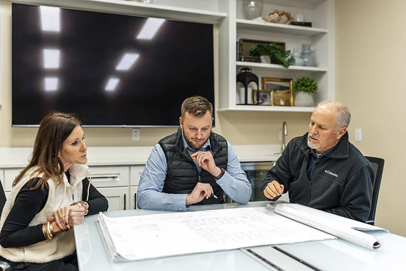 Three people sitting at a table in a meeting room, reviewing blueprints or plans, with a large TV screen behind them and shelves with decorative items and a small plant on the wall.
