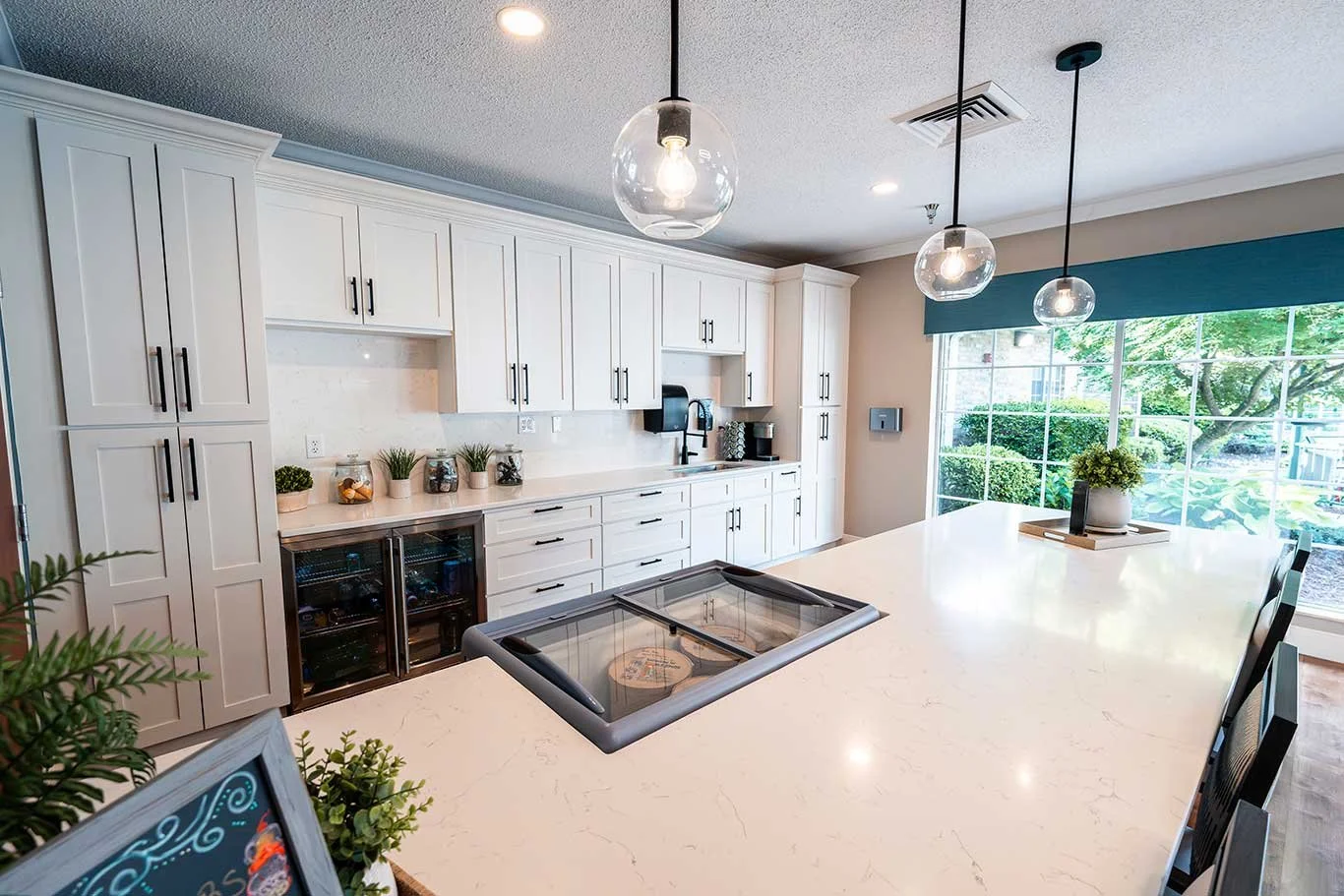 Modern kitchen with white cabinetry, large white countertop island, pendant lights, and large window with a view of greenery outside