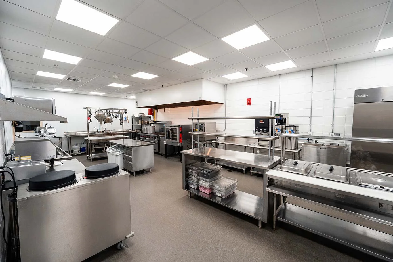 Empty commercial kitchen with stainless steel appliances, countertops, and shelves, with overhead fluorescent lighting.