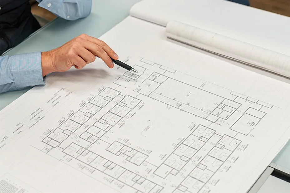 An architect or engineer's hand holding a pen pointing at a detailed building floor plan on a large sheet of paper.