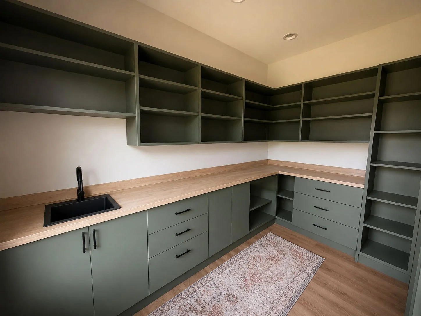 Empty modern kitchen with green cabinets, wooden countertops, black sink and faucet, open shelves, and a patterned rug.