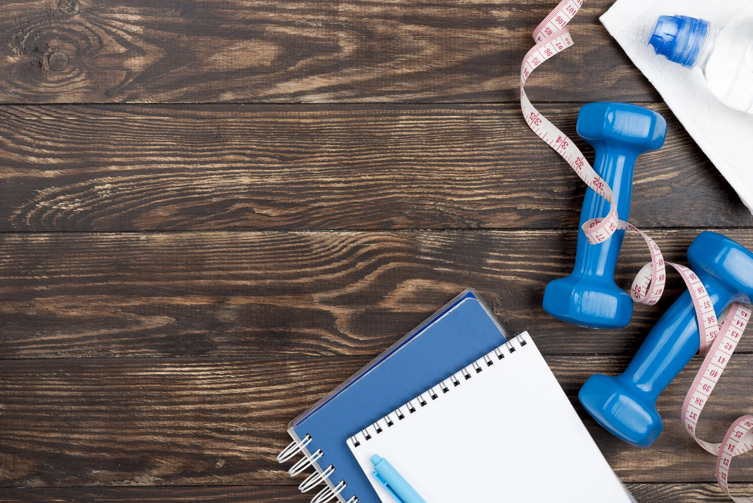 Workout accessories including blue dumbbells, a white towel, a tape measure, a blue notebook, and a pen arranged on a wooden surface with a spiral notebook.