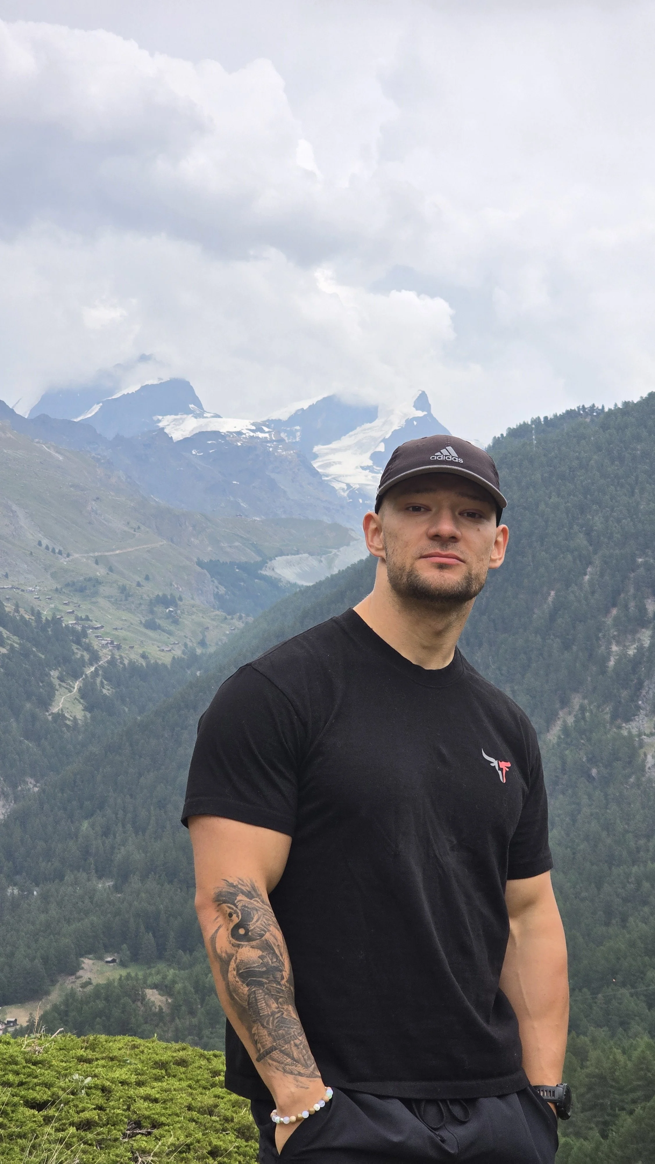 Man in black t-shirt and cap standing outdoors with mountain landscape and snow-capped peaks in the background.