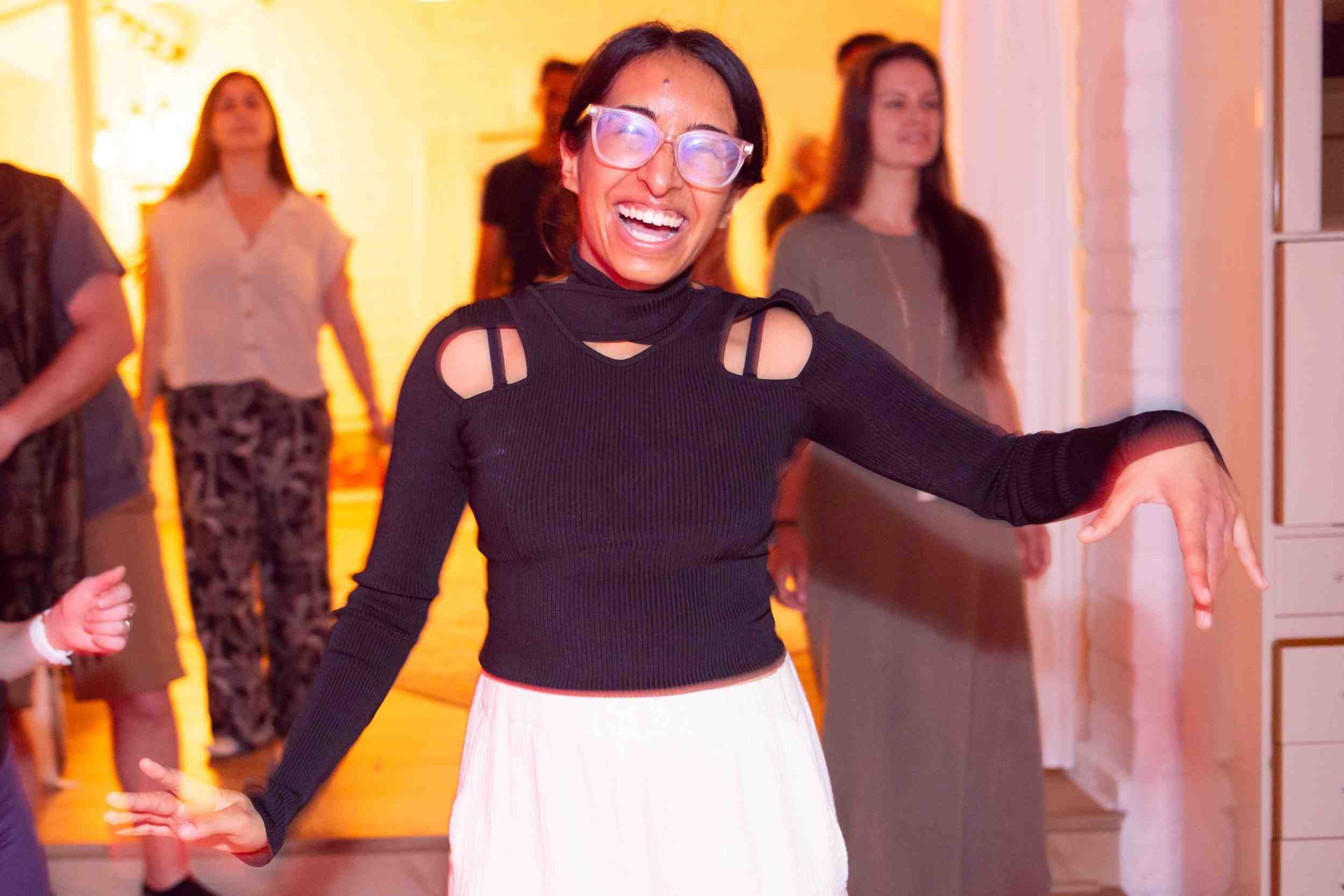 A woman with short black hair, wearing large glasses, a black cut-out shoulder top, and a white skirt, is smiling widely and appears to be dancing or enjoying herself at a social event.