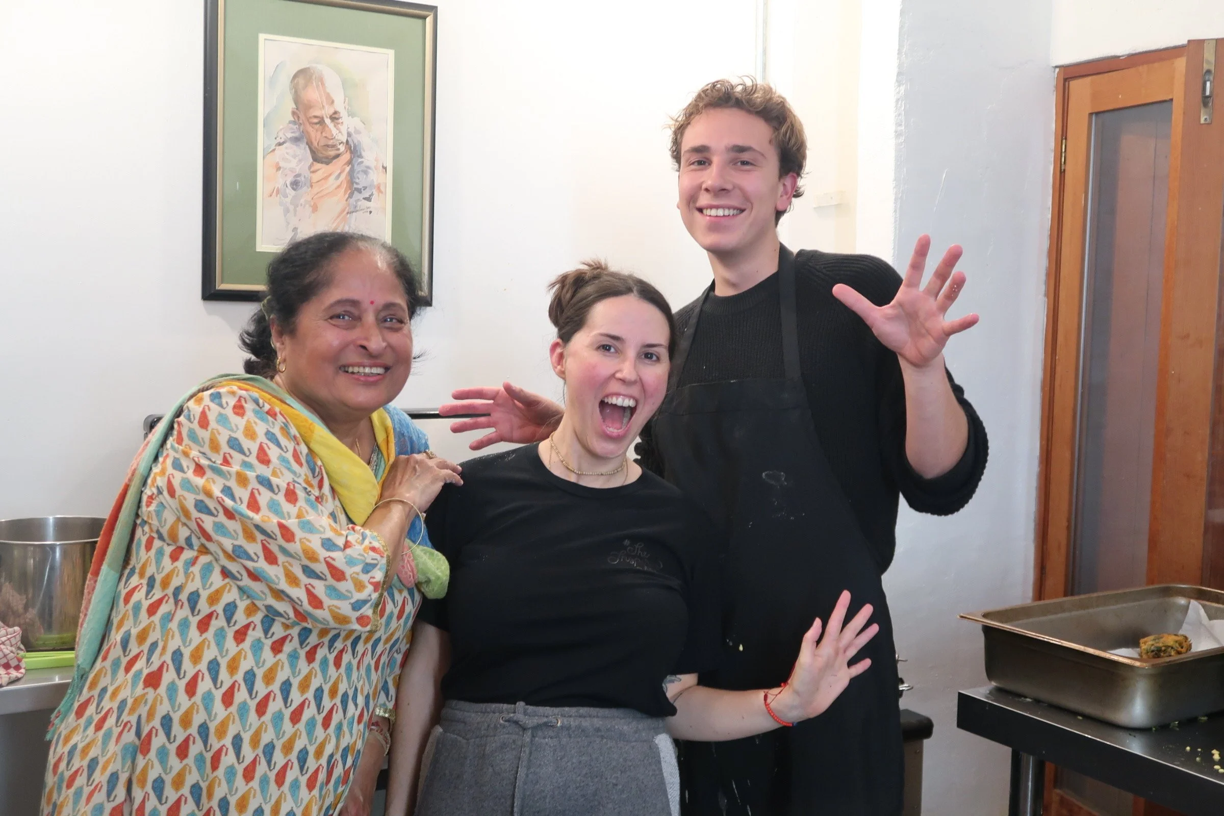 Three people smiling and posing in a kitchen. An older woman in a colorful traditional dress, a young woman with dark hair in a black shirt and jeans, and a young man with curly hair in a black shirt. There are paintings on the wall behind them, and cooking utensils and food on the counter.