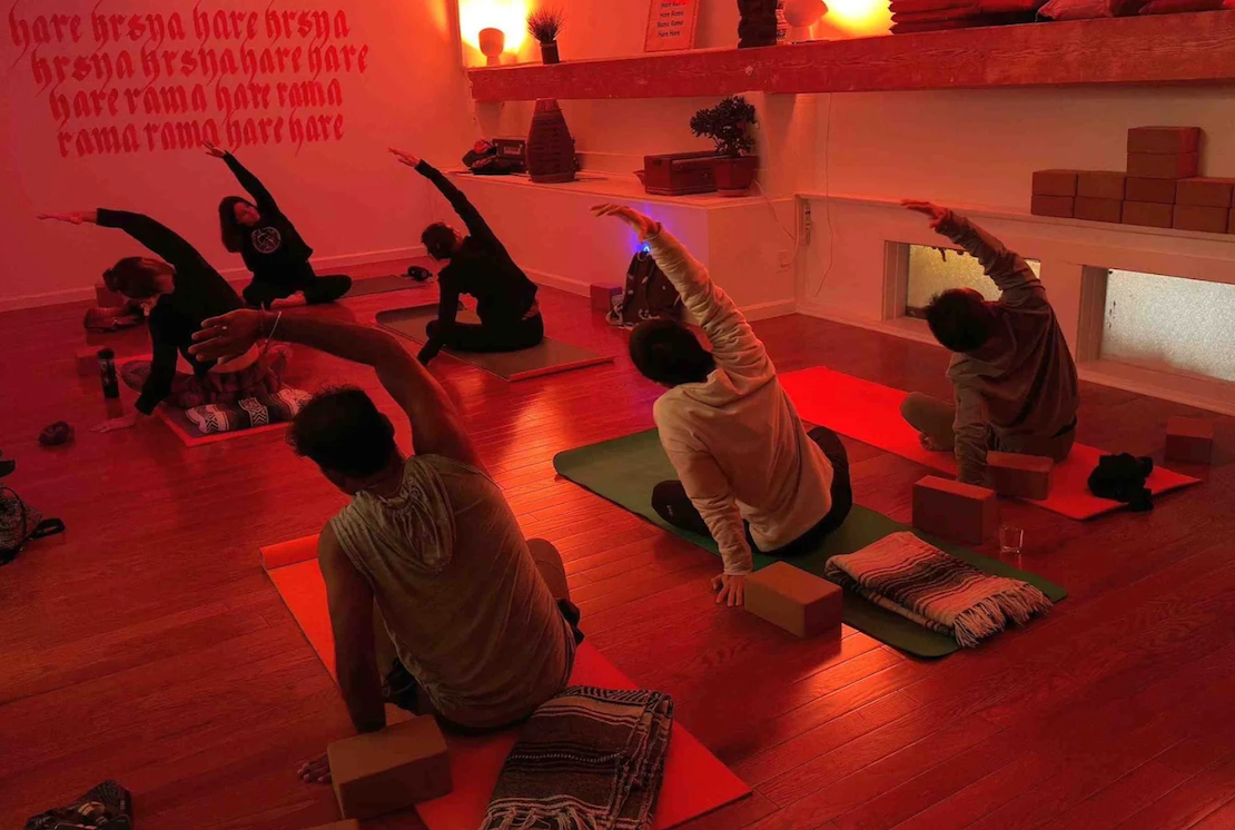 People participating in a yoga class in a room with orange lighting, practicing a side stretch pose on yoga mats with blocks, towels, and water bottles nearby.