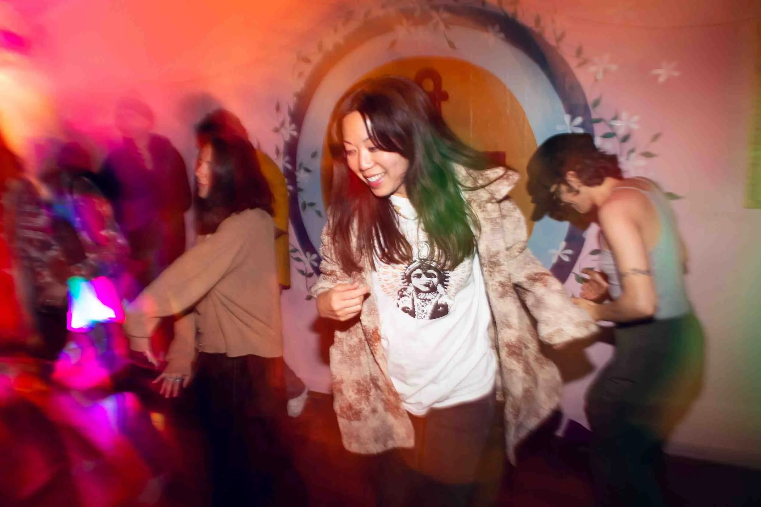 A woman dancing smiling at a party with colorful lighting and other people in the background.