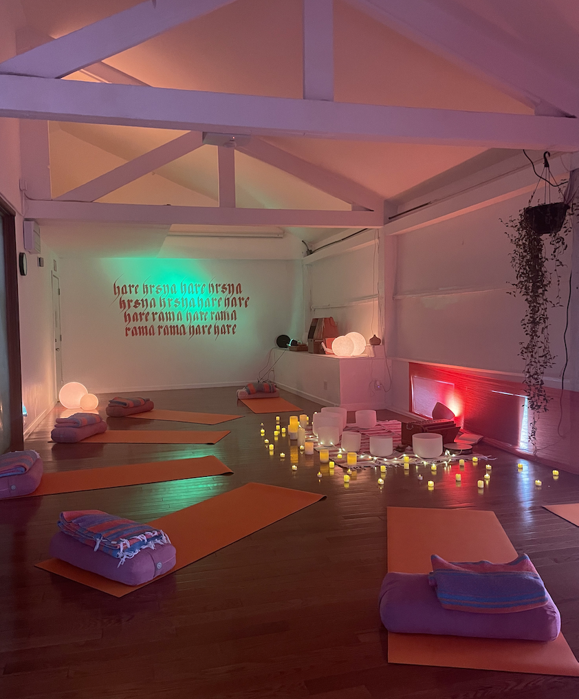 Yoga or meditation studio with orange mats and folded blankets, lit by candles and colorful lights, featuring a wall with text in red and green and hanging plants.