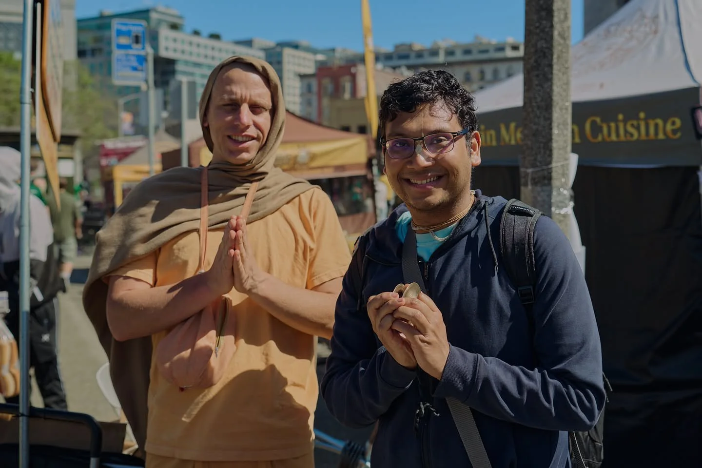 Two men smiling at a street fair, one in traditional Hindu attire and the other in casual clothing with a backpack.