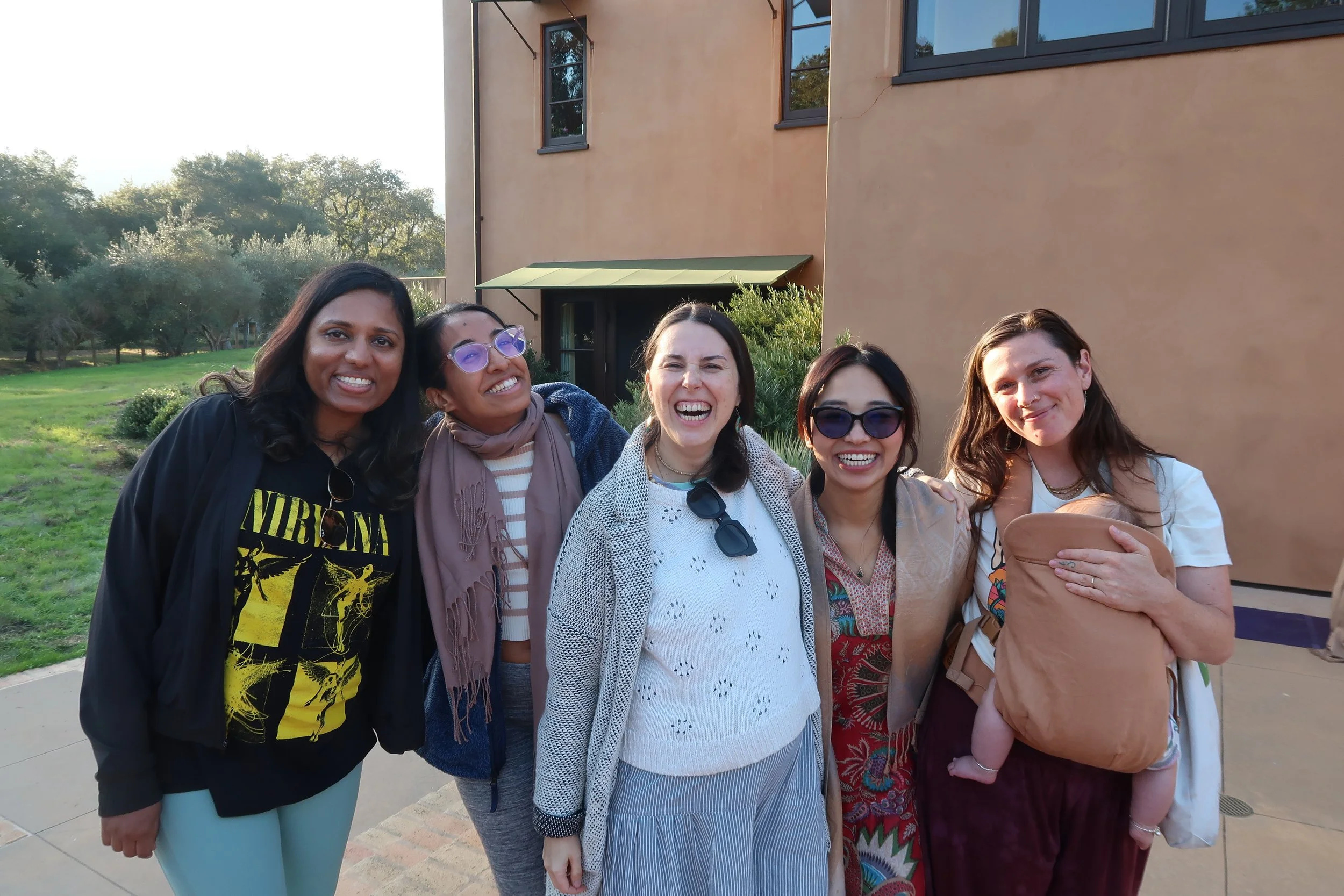 Group of five women smiling and posing outdoors in front of a beige building and trees.