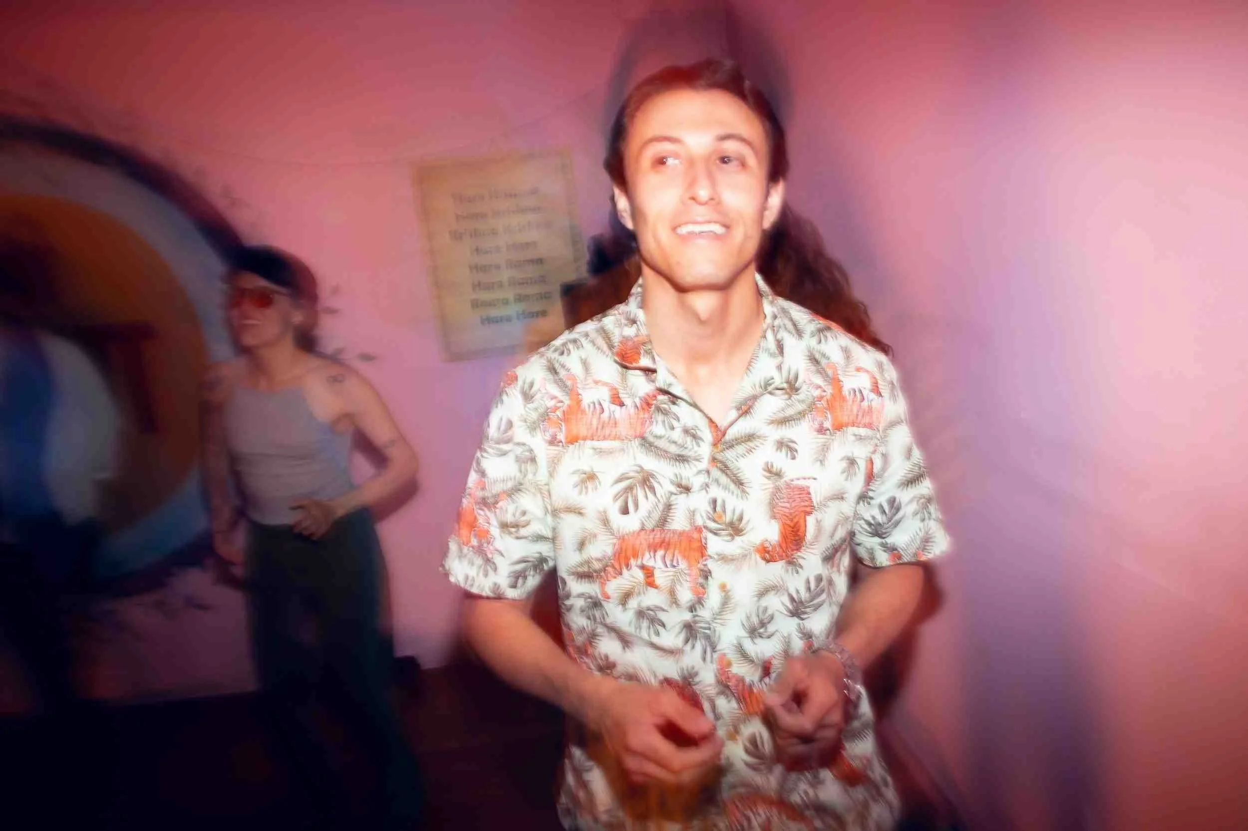 A young man with long hair wearing a colorful short-sleeve Hawaiian shirt with a tiger pattern stands against a pink wall. In the background, a woman with sunglasses, a beige top, and a tattoo on her arm stands near the wall, with a piece of paper or menu attached to the wall behind her.