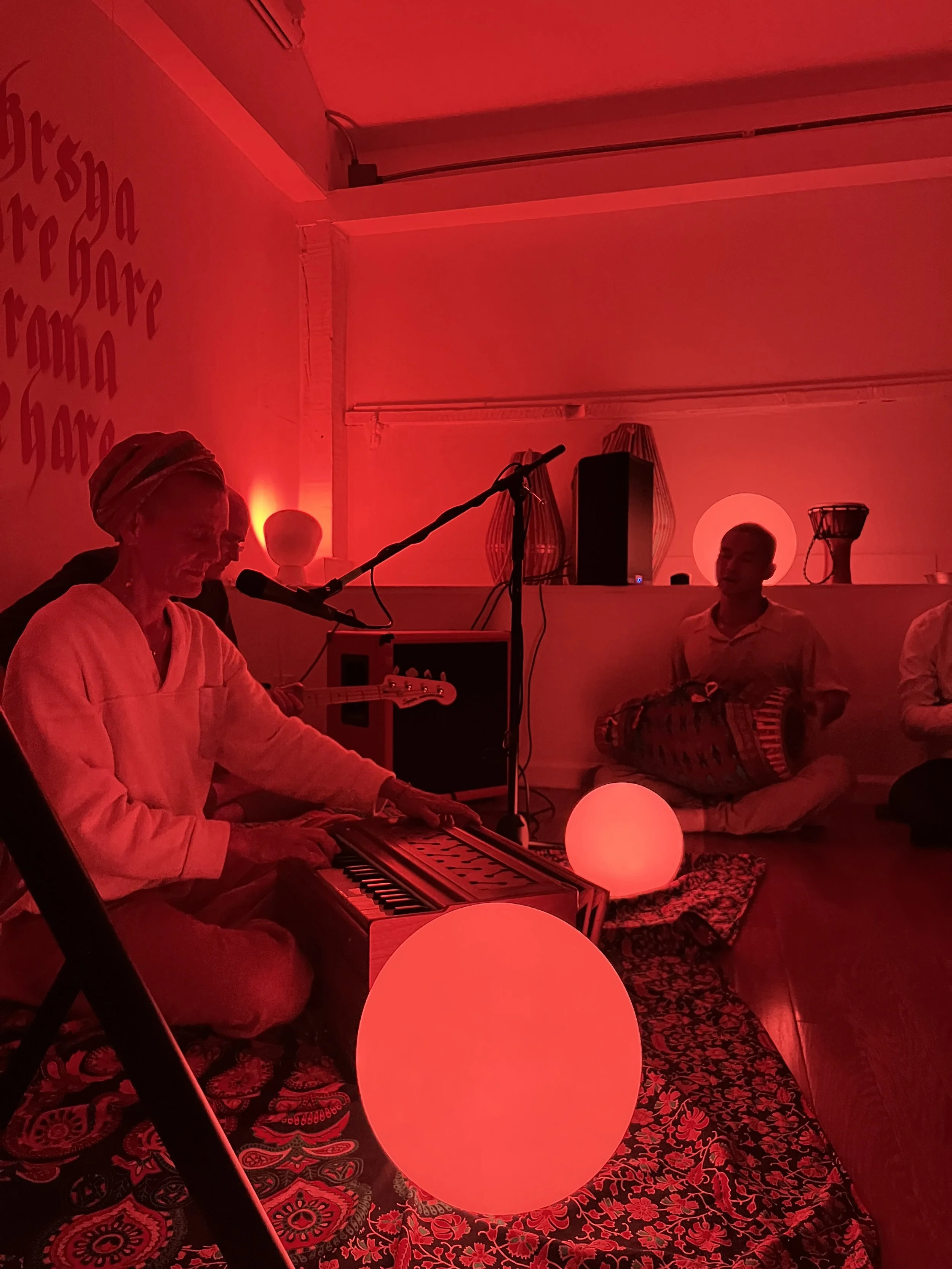 A person playing keyboard and singing into a microphone, two other people sitting and listening, a large glowing red ball on the floor, illuminated by red light, in a cozy room decorated with patterned carpet and wall art.