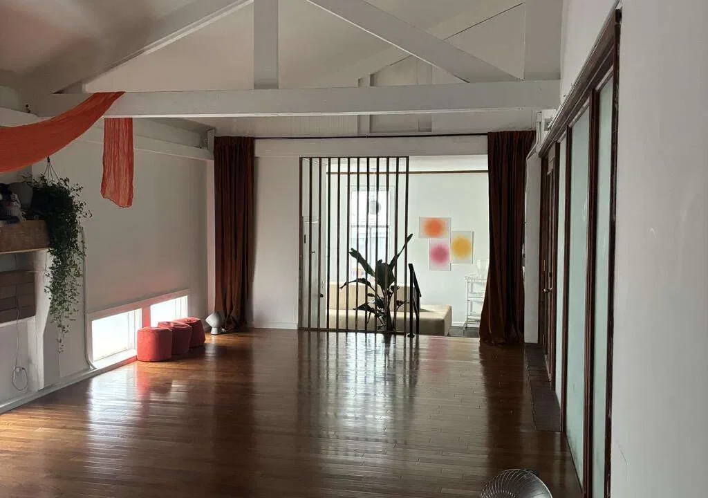 Empty room with wooden flooring, directional brown curtains, a large potted plant near the staircase, colorful abstract art on the wall, and a small window near the floor with pink poufs underneath.