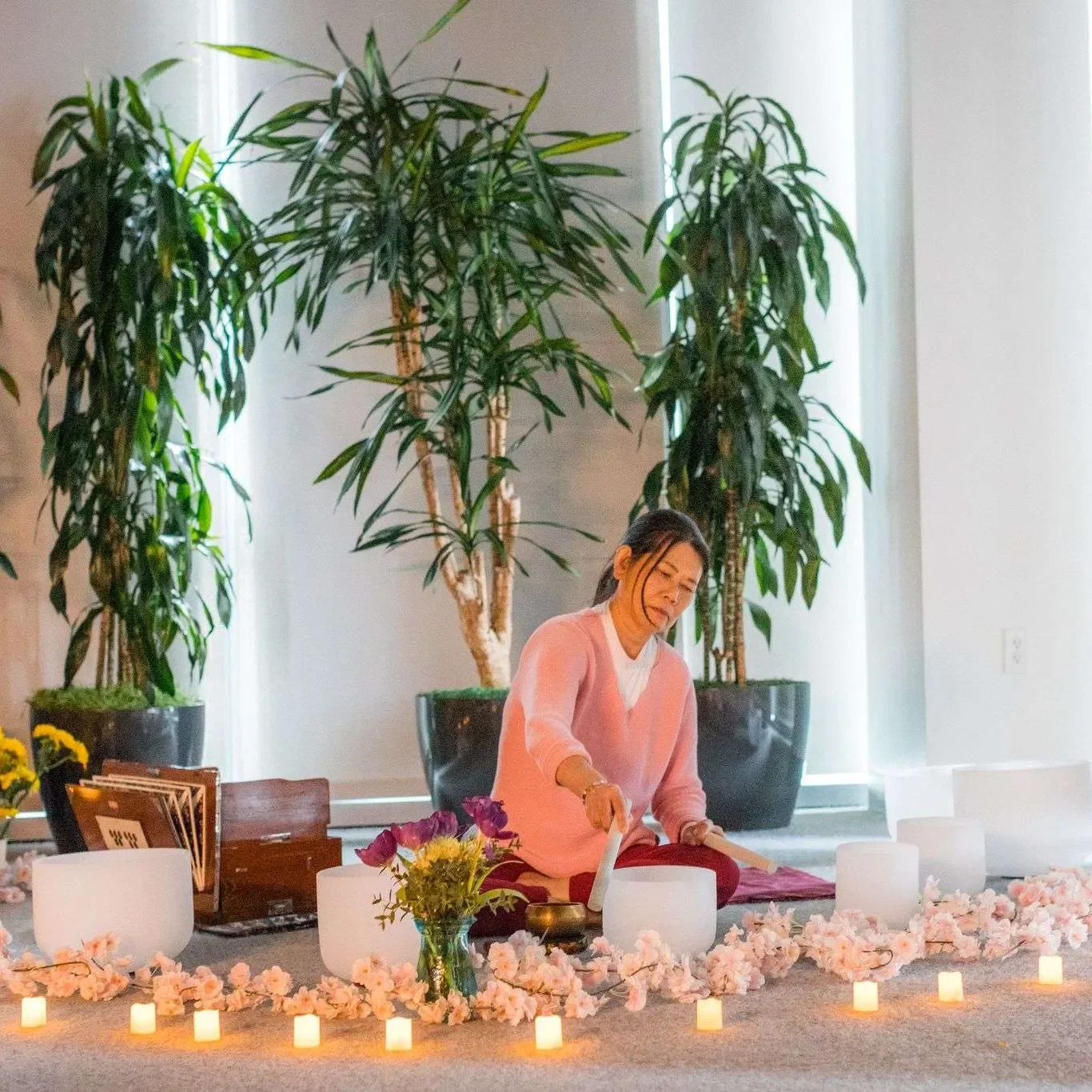 A woman in a pink sweater sitting on the floor surrounded by pink and white flowers and candles, with tall green plants behind her, engaged in a meditation or ritual.