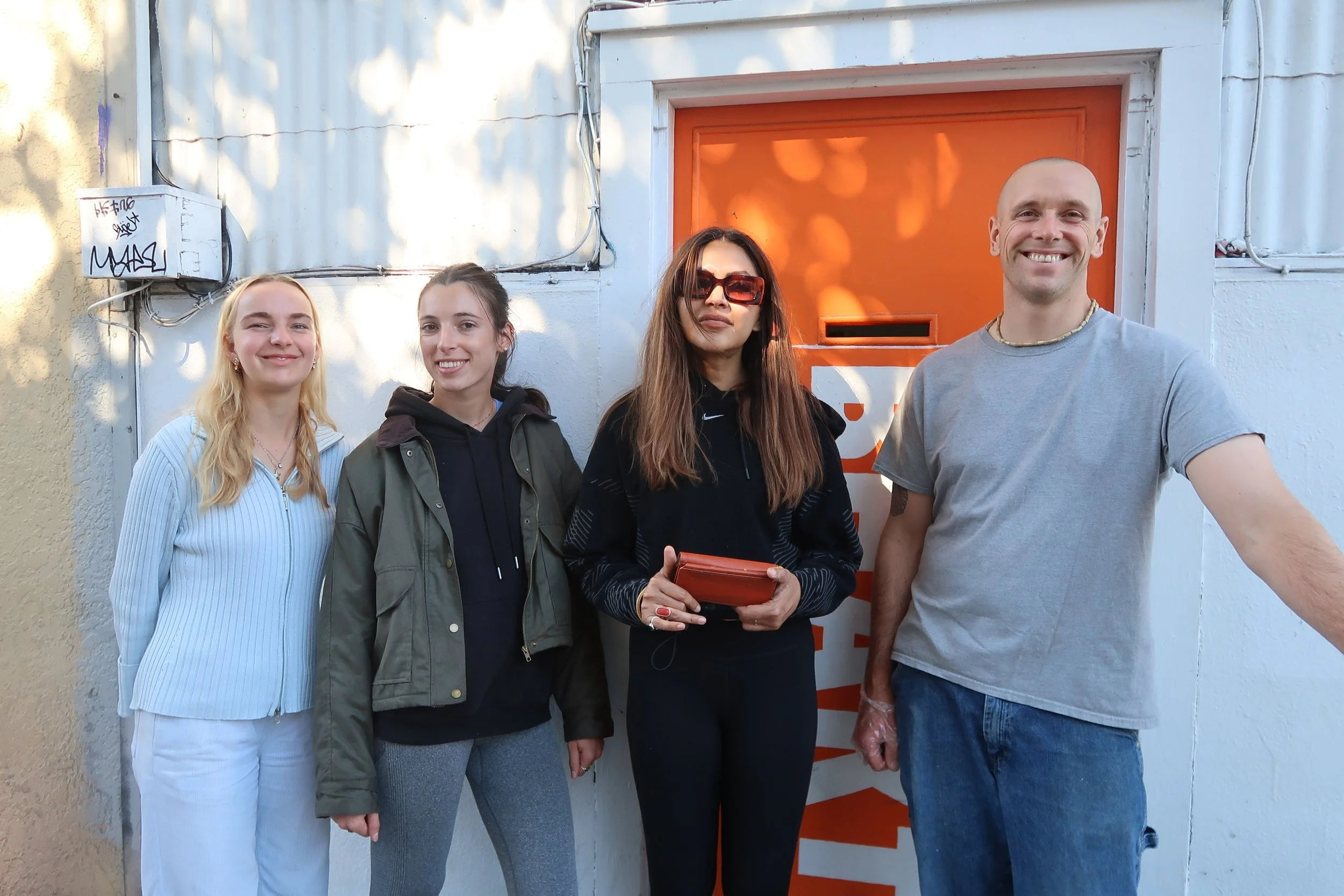 Four people standing outdoors in front of an orange door. From left to right: a blonde woman in a light blue and white striped zip-up top, a woman with brown hair in a dark hoodie and green jacket, a woman with long brown hair wearing large sunglasse