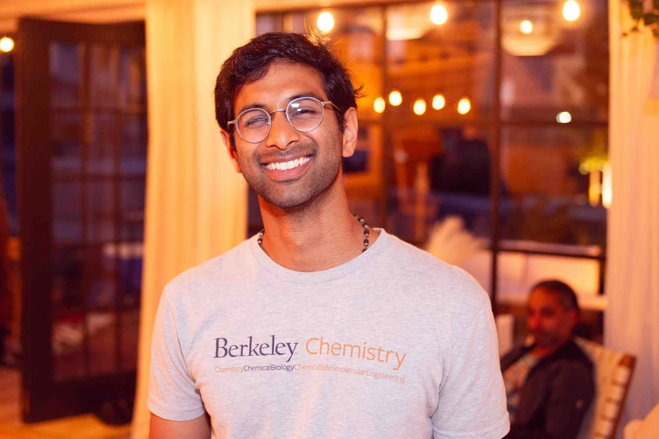 A man with glasses smiling at a gathering, wearing a Berkeley Chemistry T-shirt.