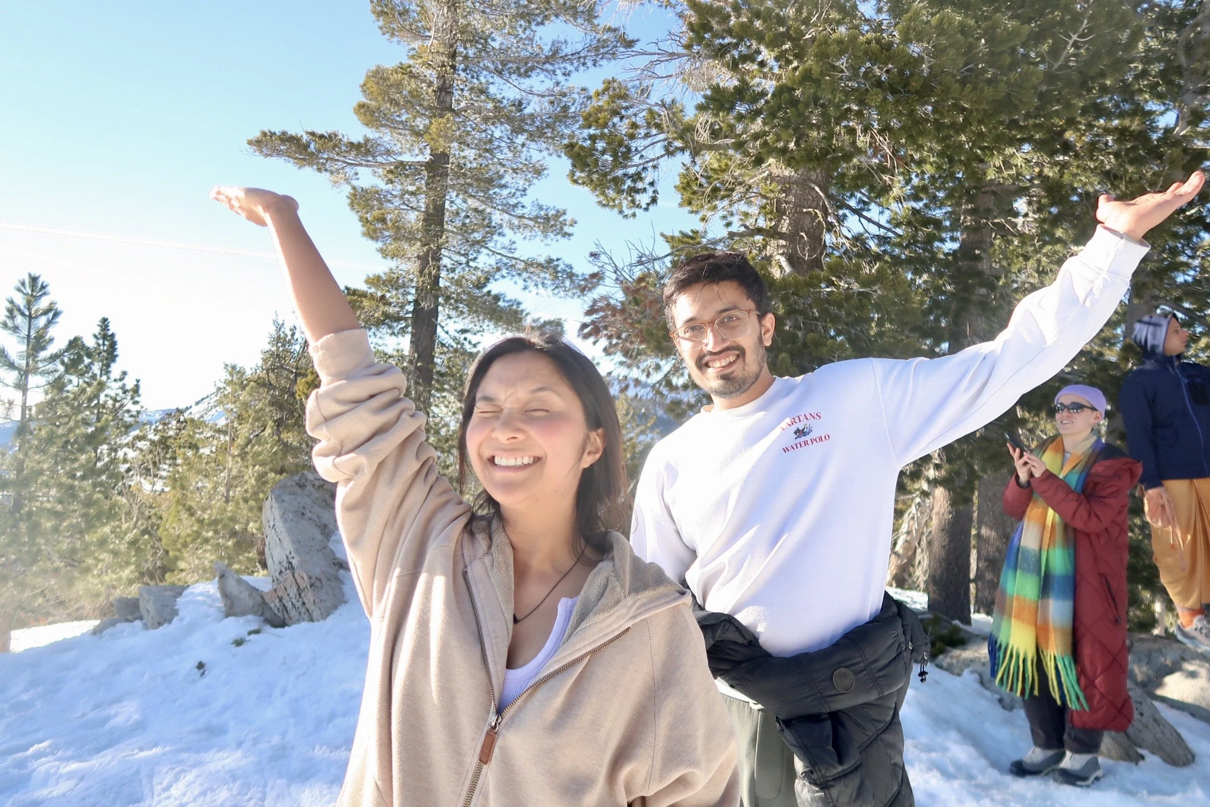 A group of people outdoors in a snowy forest, smiling and celebrating, with one man and woman in the foreground, and others in the background, surrounded by tall pine trees and a bright blue sky.