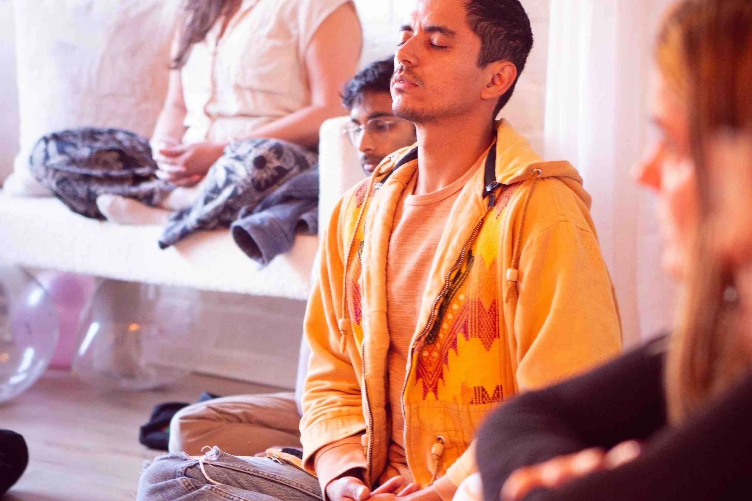 Group of young people sitting on the floor, some with eyes closed, possibly meditating or relaxing in a calm setting.