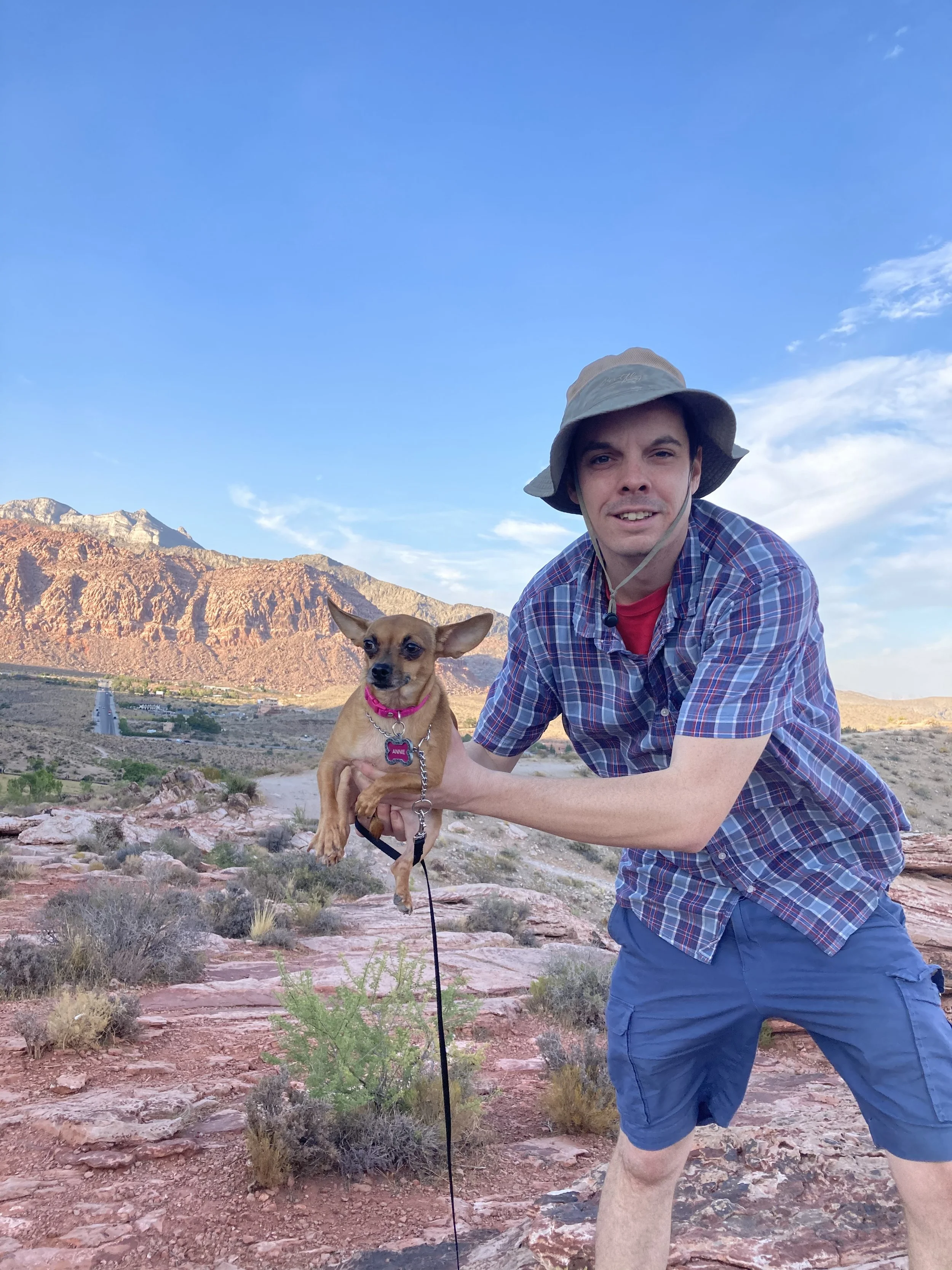 A man crouching in a desert landscape with a small dog, mountains in the background, and a clear blue sky.
