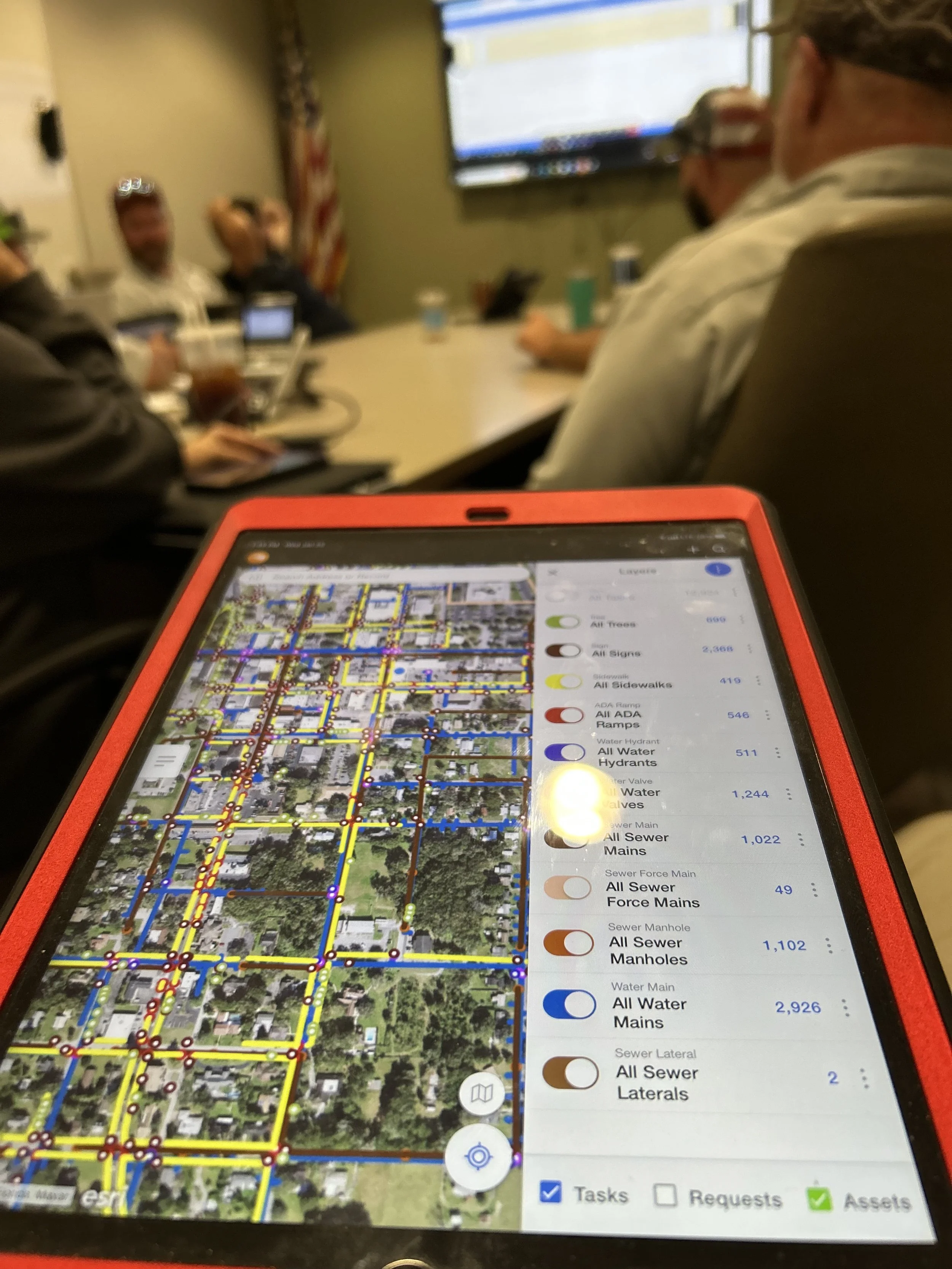 A tablet displaying a detailed map of a city's water and sewer infrastructure with color-coded lines and features for different utilities. The field crew is blurred in the background attending a meeting.