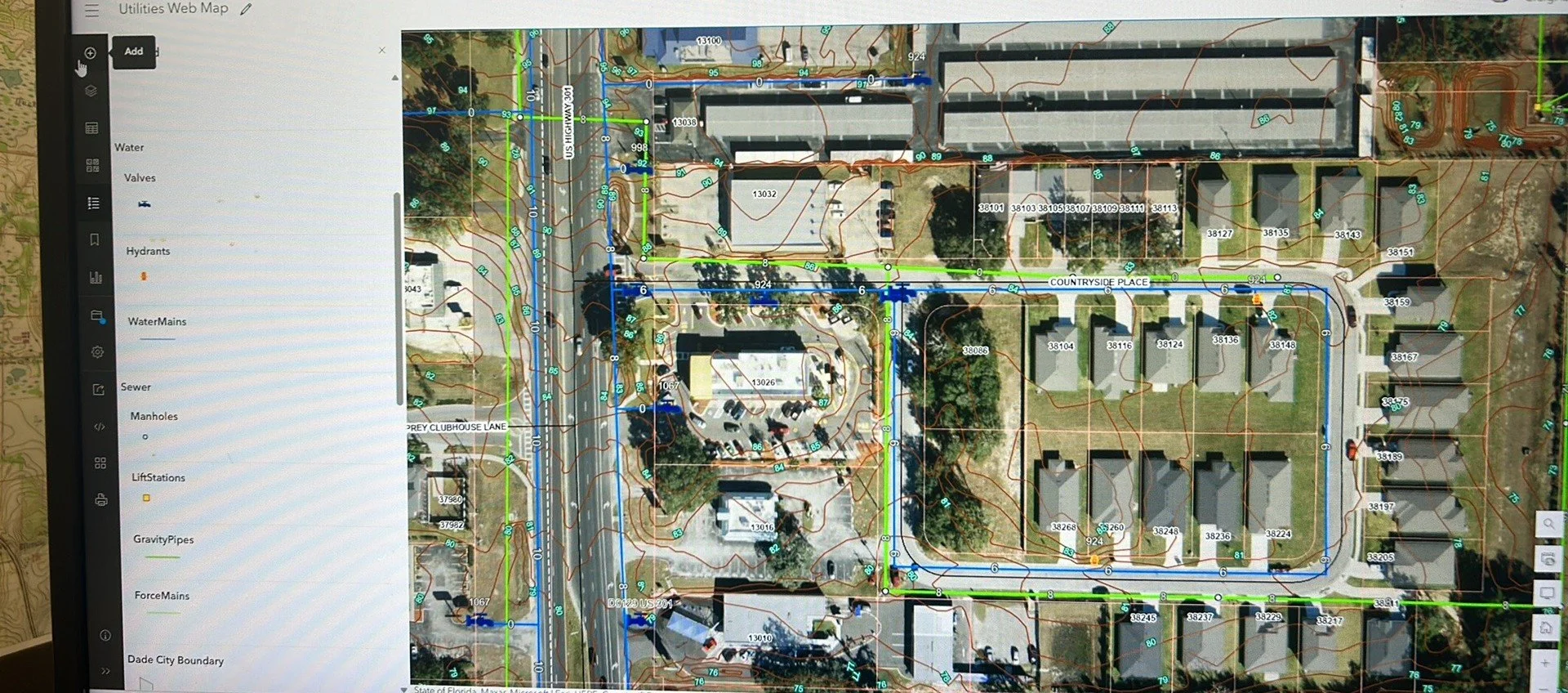 A detailed map of a residential area showing water lines, valves, hydrants, manholes, lift stations, pressure pipes, sewage, and other infrastructure, overlaid on an aerial photo of houses, streets, and greenery.